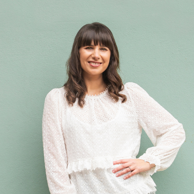 Headshot of author Gab Tozer with shoulder-length brown hair and bangs, smiling against a light green wall. She is wearing a white eyelet lace blouse and has one hand resting on her hip.