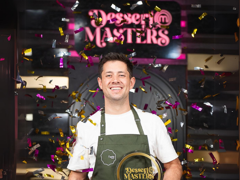 Image of Gareth Whitton standing in the Dessert Masters kitchen. He has a big smile on his face. Large confetti tinsle is falling around him and he is holding a dessert Masters trophy.