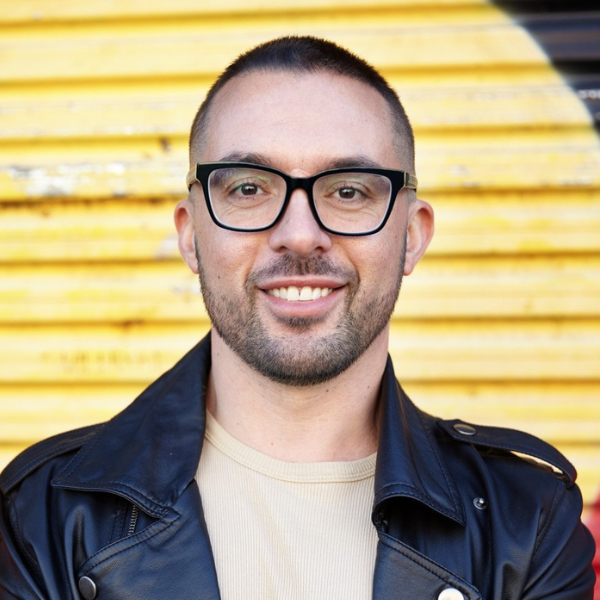 Image of artist, Mitch Hibbens. Photo is a Headshot image of them standing in front of a large mural of the Aboriginal Flag. They are mearing a black leather jacket and light beige t-shirt. They are wearing black rimmed glasses and are staring straight at the camera. They are smiling.