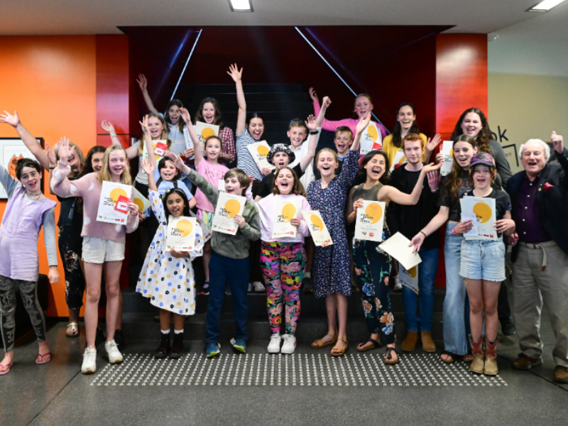 A large number of joyous young people standing in front, and on the stairs at the Library Museum. They are all holding there winner's certificates in the air.