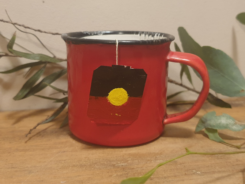 image of red metal mug with tea bag label hanging over the edge. The label has the Aboriginal flag on it. The mug is sitting on a shelf amongst Eucalypt leaves.