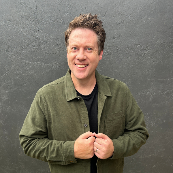 Headshot of author Adrian Beck with short brown hair, smiling in front of a dark grey wall. He is wearing an olive green button-down jacket over a black t-shirt.