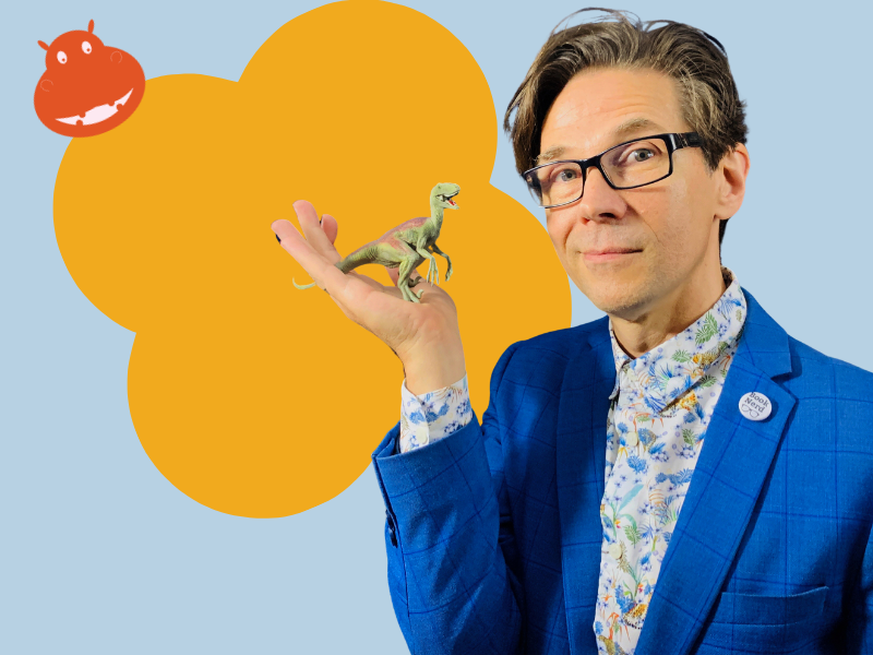 Image of Author George Ivanoff. He is blue suit with white and blue floral shirt. He is holding his hand palm upwards with a two dinosaur on it.