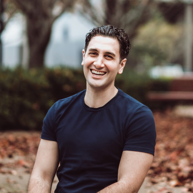 Headshot of author Will Kostakis with short dark hair, laughing warmly. He is wearing a dark blue t-shirt, seated outdoors with a soft-focus background of trees and fallen leaves.